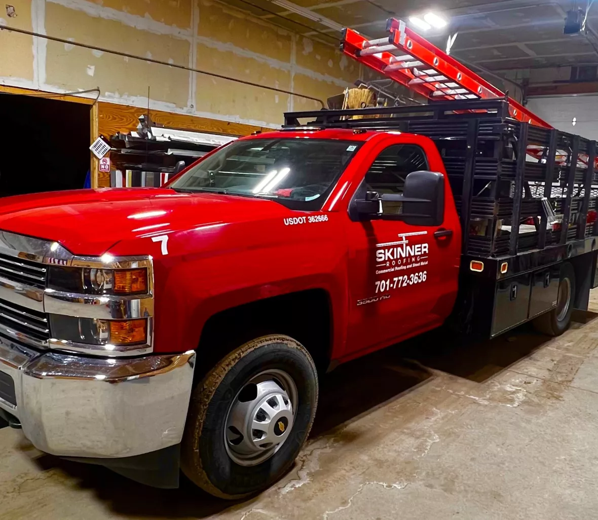 A red Chevrolet pickup truck with a black rack, displaying 'Skinner Roofing' logo and a ladder on top, parked indoors.