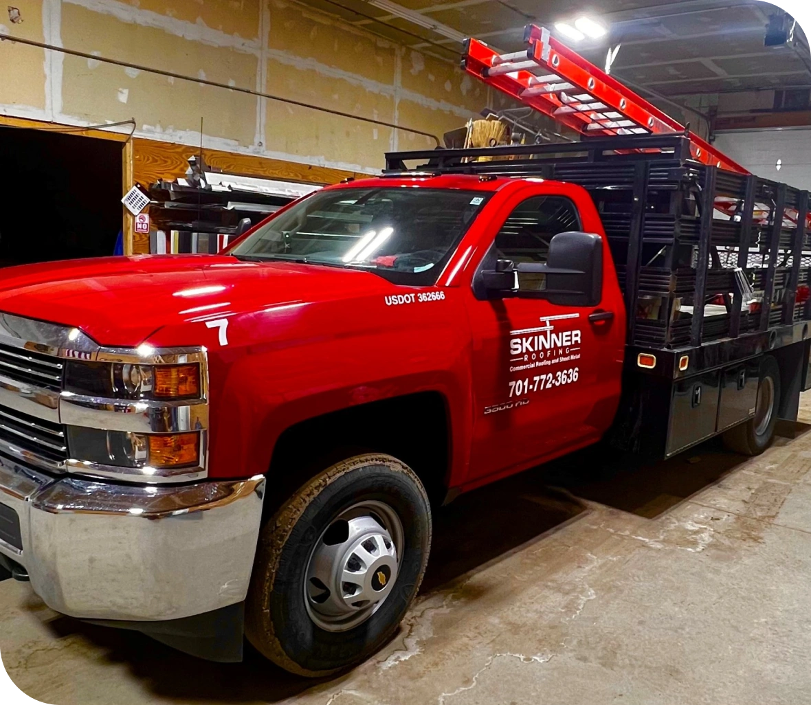 A red Chevrolet pickup truck with a black rack, displaying 'Skinner Roofing' logo and a ladder on top, parked indoors.